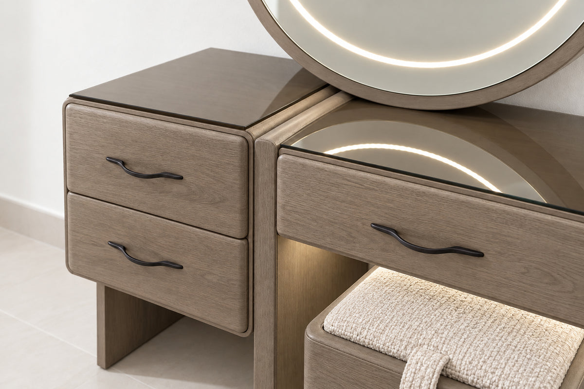 A modern vanity table with two drawers, a dark brown top, and a matching upholstered side panel, shown alongside a lit round mirror and a textured stool cushion.