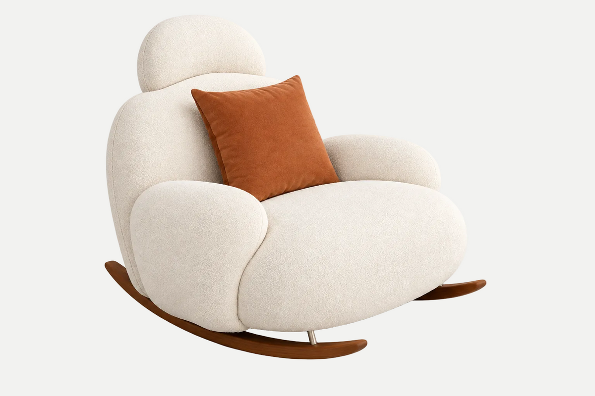 A modern rounded rocking chair upholstered in warm off-white fabric, featuring a contrasting rust-orange throw pillow and dark brown wooden rockers, set against a clean white background.