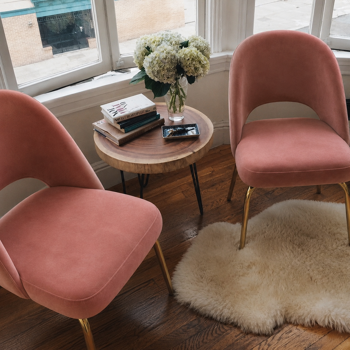 Two blush pink velvet armchairs with gold legs in a bright room, with a small round wood table in between them holding books and a vase of flowers.