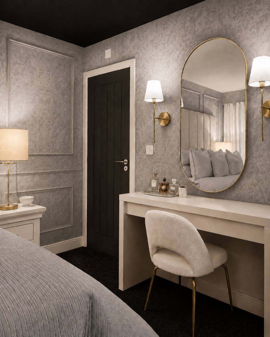 A styled bedroom interior featuring a white Accent upholstered Chair and white desk with a matching a large oval gold-framed mirror, wall sconces, textured gray wallpaper, and a dark wood door. A bed is partially visible in the foreground.
