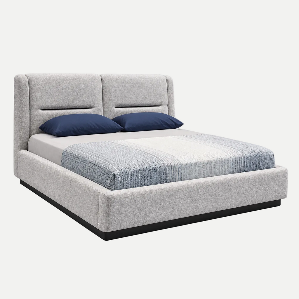 A modern platform bed with a light gray upholstered frame and tall segmented headboard, styled with a striped blue and white duvet and two navy blue pillows, shown on a light gray background.