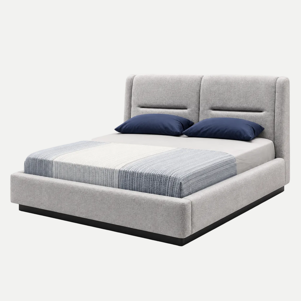 A modern platform bed with a light gray upholstered headboard and base, styled with a striped blue and white comforter and two navy blue pillows, set against a plain white background.