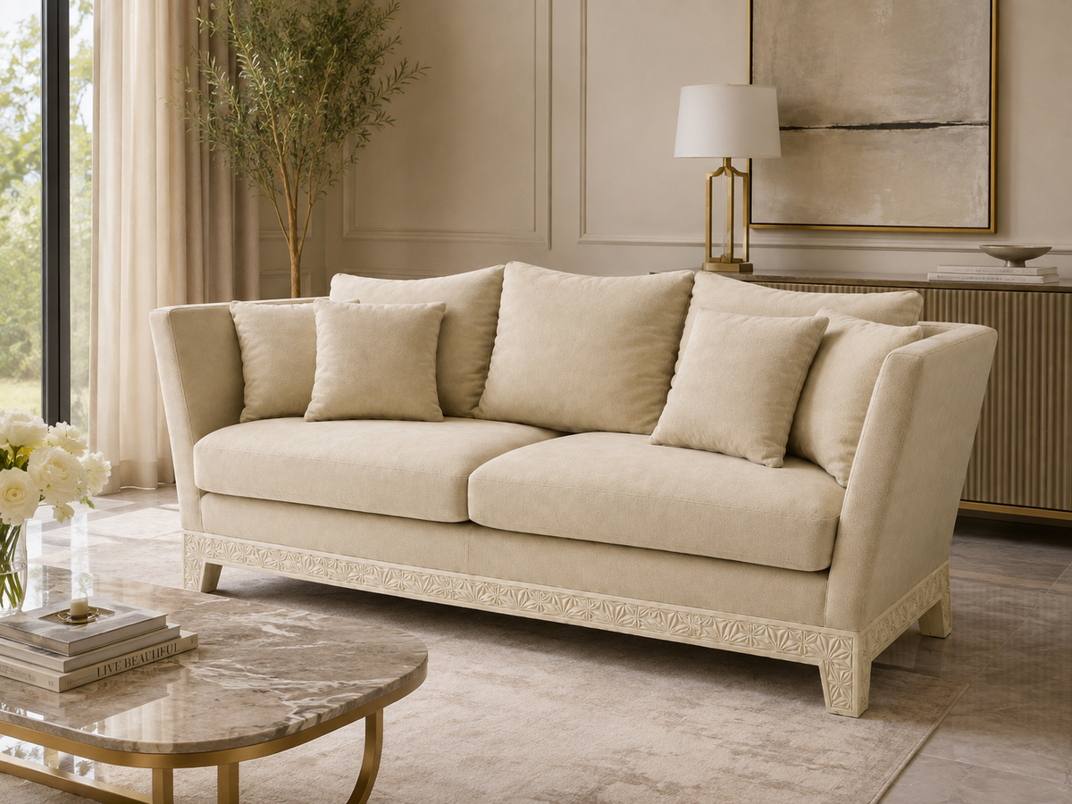 A cream-colored sofa with matching throw pillows, set in a beautifully decorated living room with beige walls, a marble coffee table, and a lamp in the background.