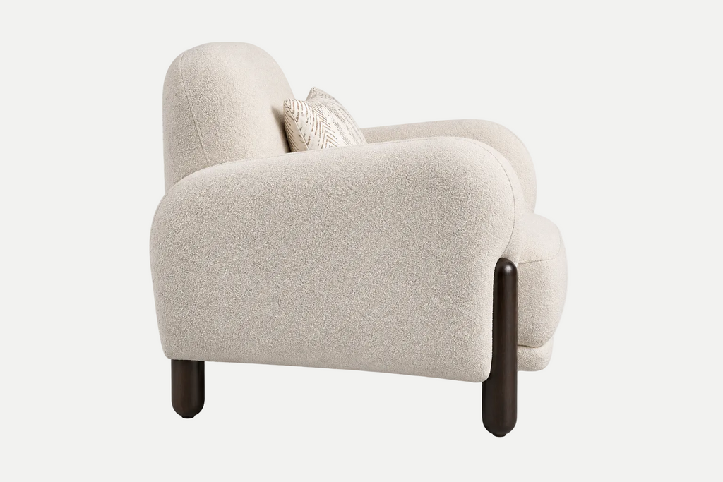 A modern low-profile armchair upholstered in a textured cream bouclé fabric, featuring dark brown tapered wooden legs and a small textured cream throw pillow, shown against a plain white studio background.