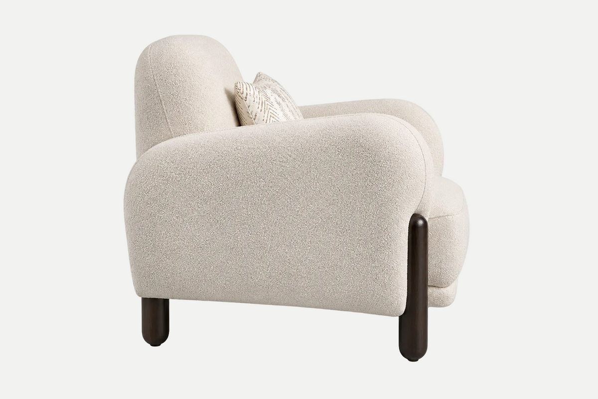 A modern low-profile armchair upholstered in a textured cream bouclé fabric, featuring dark brown tapered wooden legs and a small textured cream throw pillow, shown against a plain white studio background.