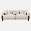 A modern two-seater sofa upholstered in a light beige textured fabric, featuring dark brown cylindrical legs and four decorative throw pillows, shown against a plain light gray background.