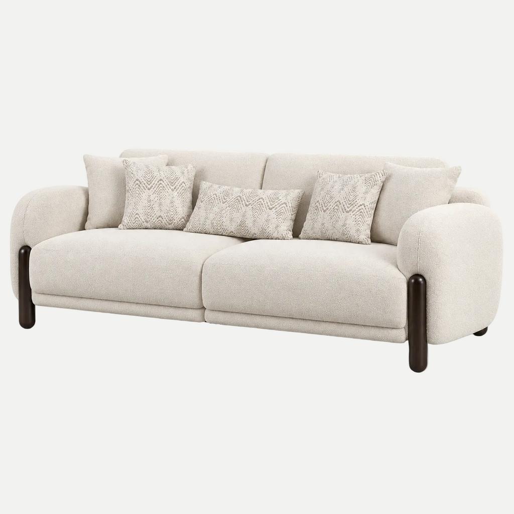 A modern three-seater sofa upholstered in a light beige textured fabric, featuring dark brown wooden legs and four decorative throw pillows with a textured geometric pattern, shown against a plain light gray background.