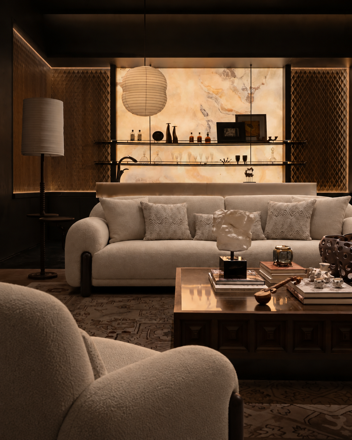 A luxury modern living room featuring a cream-colored sofa with textured throw pillows, a dark wood coffee table, and a warm, ambient background with a marble wall and decorative shelves.