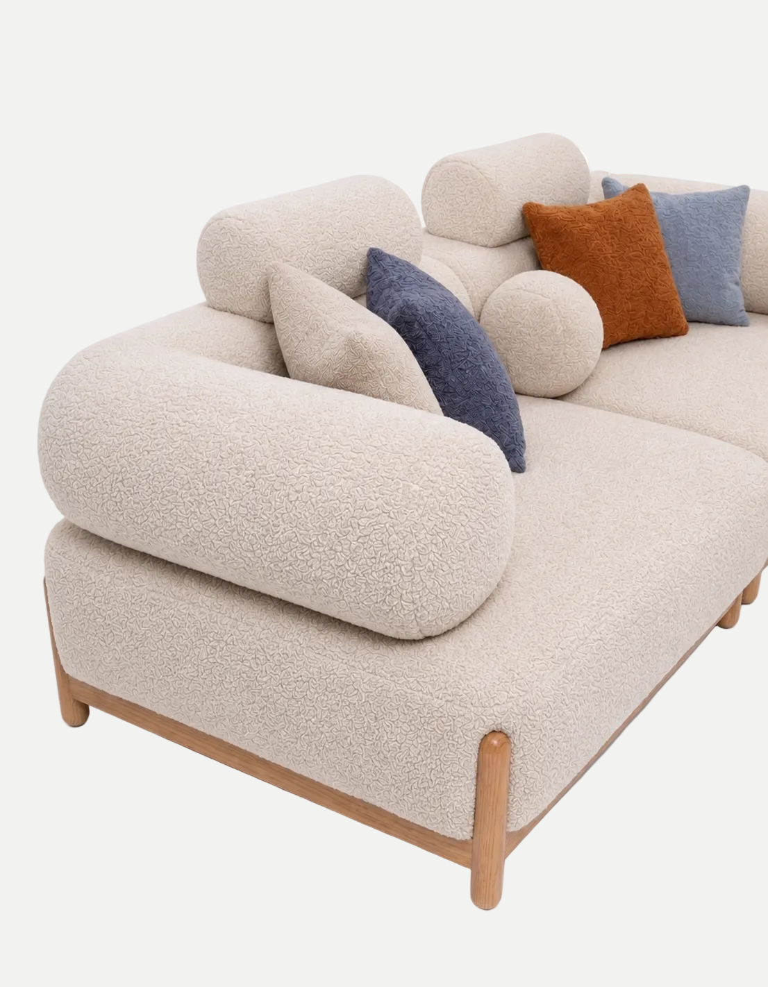 A modern curved sofa upholstered in textured cream bouclé fabric with light natural wood legs, styled with several accent pillows in blue, rust orange, and light gray on a white background.