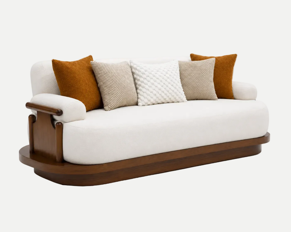 A modern three-seat sofa with a cream-colored upholstered cushion, a curved dark brown wooden base and armrests, and four decorative throw pillows in textured beige, white, and rust orange, set against a plain white background.