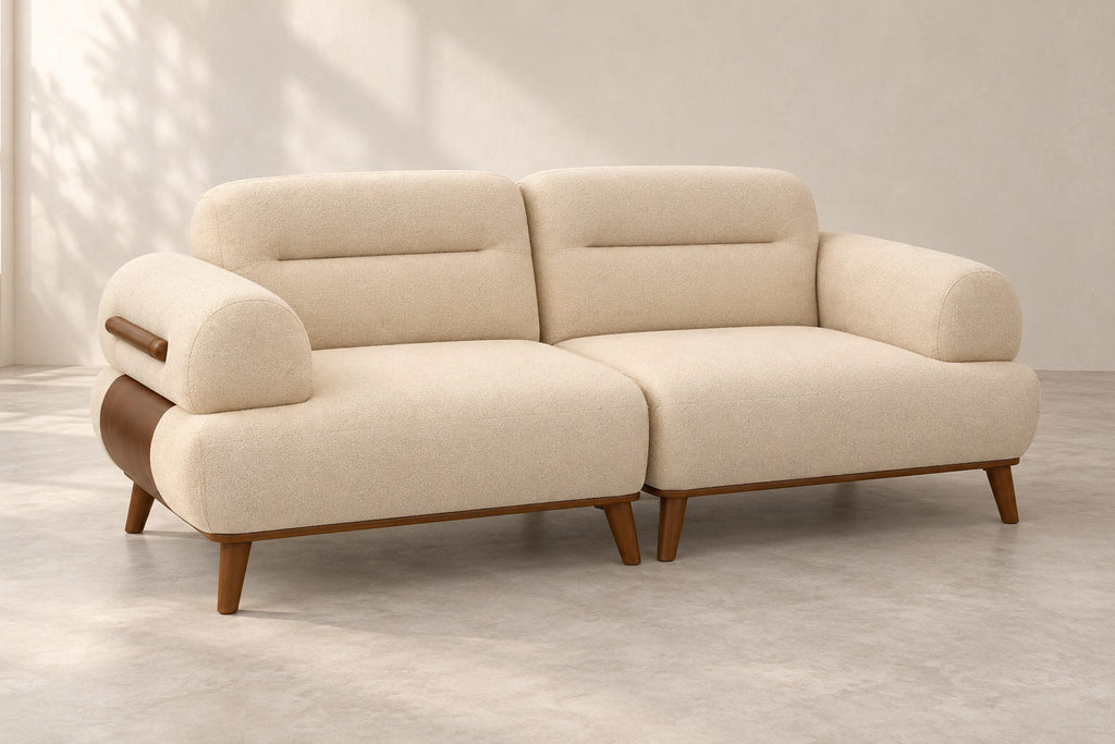 A modern two-seater sofa with cream-colored textured fabric upholstery and medium brown wooden legs, shown in a minimalist studio setting with soft window shadow patterns.
