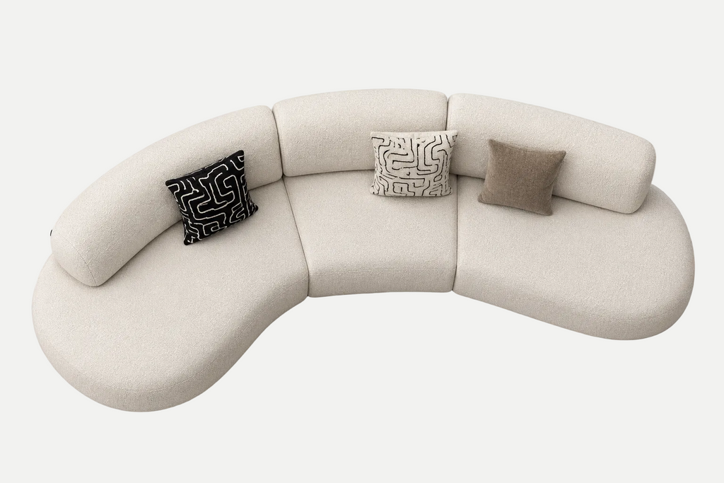 A curved modular upholstered sofa in a light warm off-white fabric, styled with three decorative throw pillows on a clean light gray background.