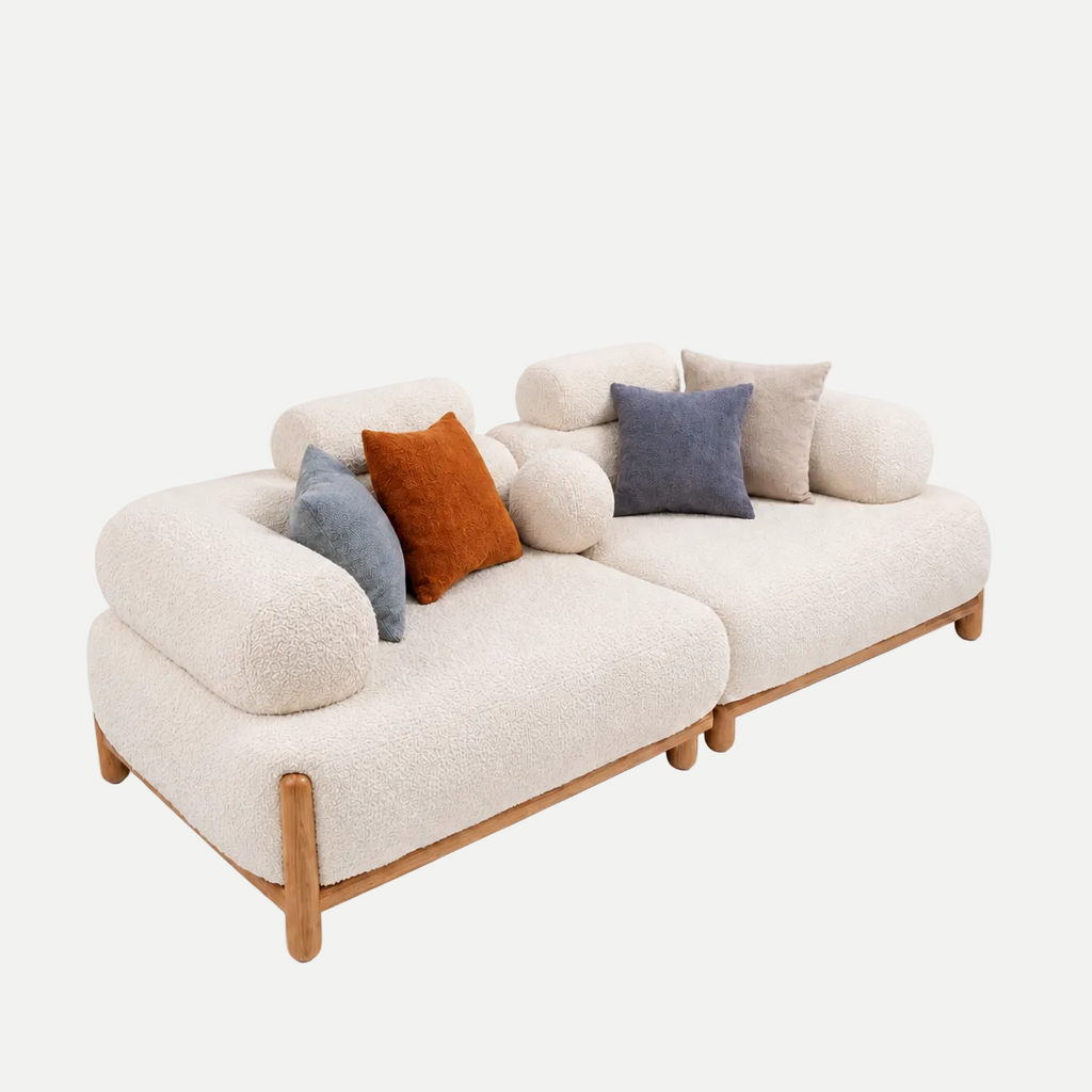 A modern two-seater sofa upholstered in cream bouclé fabric with light natural wood legs and trim, styled with several decorative throw pillows in blue, rust orange, and beige on a plain white background.