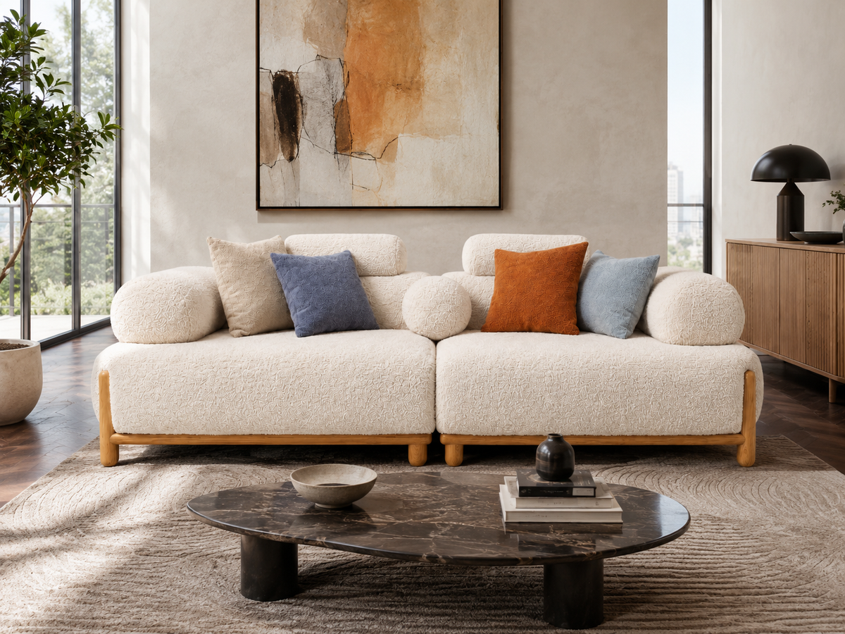 A modern two seater sofa upholstered in cream bouclé fabric with light wood legs, styled in a bright living room with cream, blue, and orange accent pillows. A dark marble coffee table and a textured rug are in the foreground, with a large abstract painting on the wall behind.