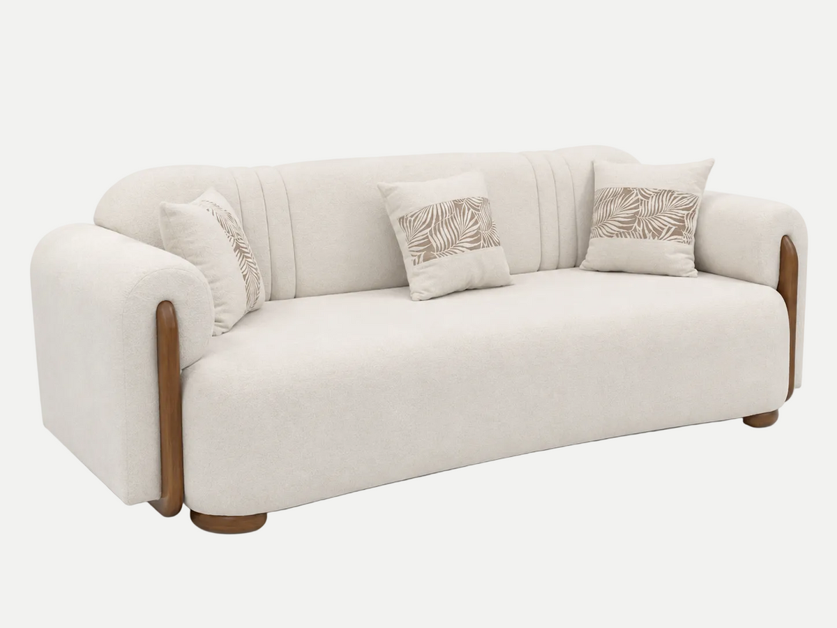 A modern three-seater sofa upholstered in cream-colored fabric with light brown wooden legs and arm accents, decorated with three matching throw pillows featuring a brown palm leaf pattern, set against a plain white background.