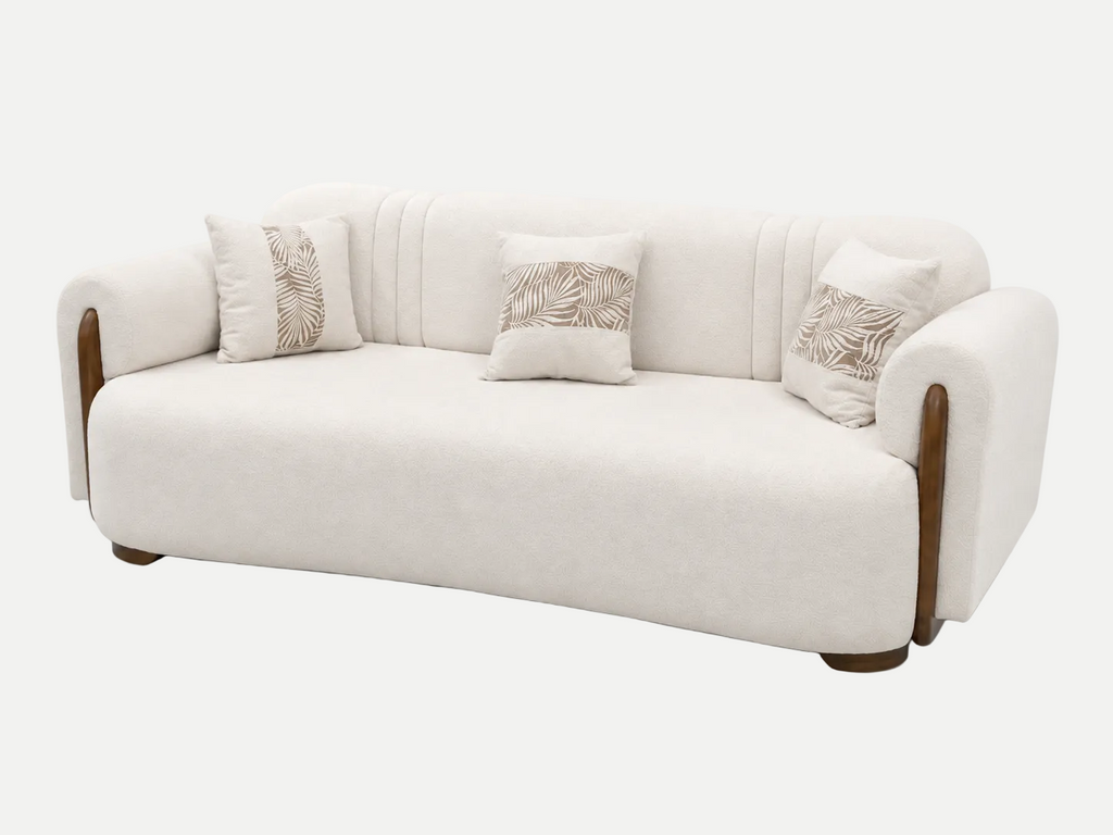 A modern curved sofa upholstered in cream-colored fabric with dark brown wooden arm accents and three matching decorative throw pillows featuring a beige leaf pattern, set against a plain white background.