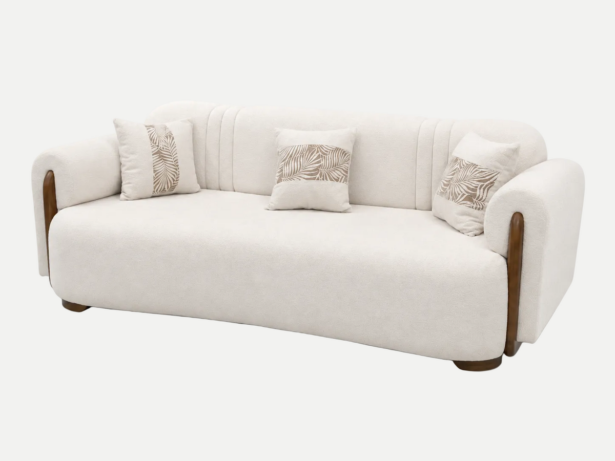 A modern curved sofa upholstered in cream-colored fabric with dark brown wooden arm accents and three matching decorative throw pillows featuring a beige leaf pattern, set against a plain white background.