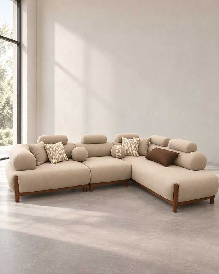 A modern sectional sofa in a warm beige fabric with dark brown wooden legs, styled with several decorative throw pillows in a minimalist, sunlit room with a large window.