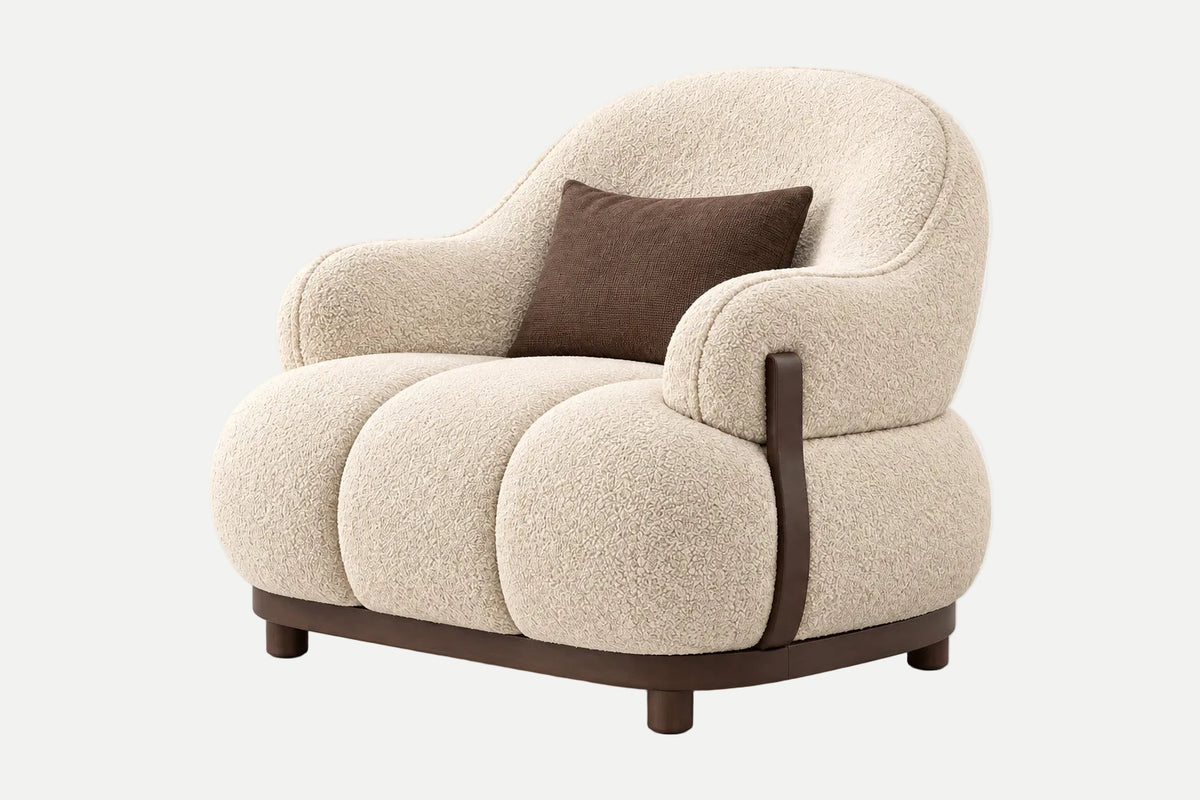 A modern rounded armchair upholstered in textured cream bouclé fabric, featuring dark brown wooden legs and a vertical armrest trim, with a matching brown accent pillow, shown on a light gray background.