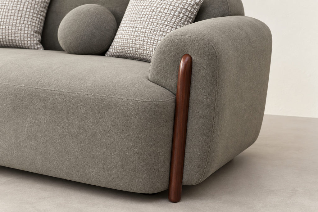 Close-up of a modern gray upholstered sofa corner with a curved wooden leg and two patterned throw pillows on a light neutral floor and wall.
