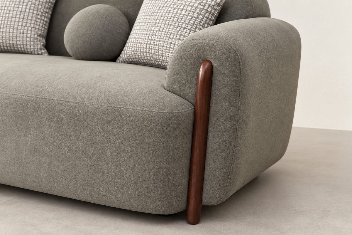 Close-up of a modern gray upholstered sofa corner with a curved wooden leg and two patterned throw pillows on a light neutral floor and wall.
