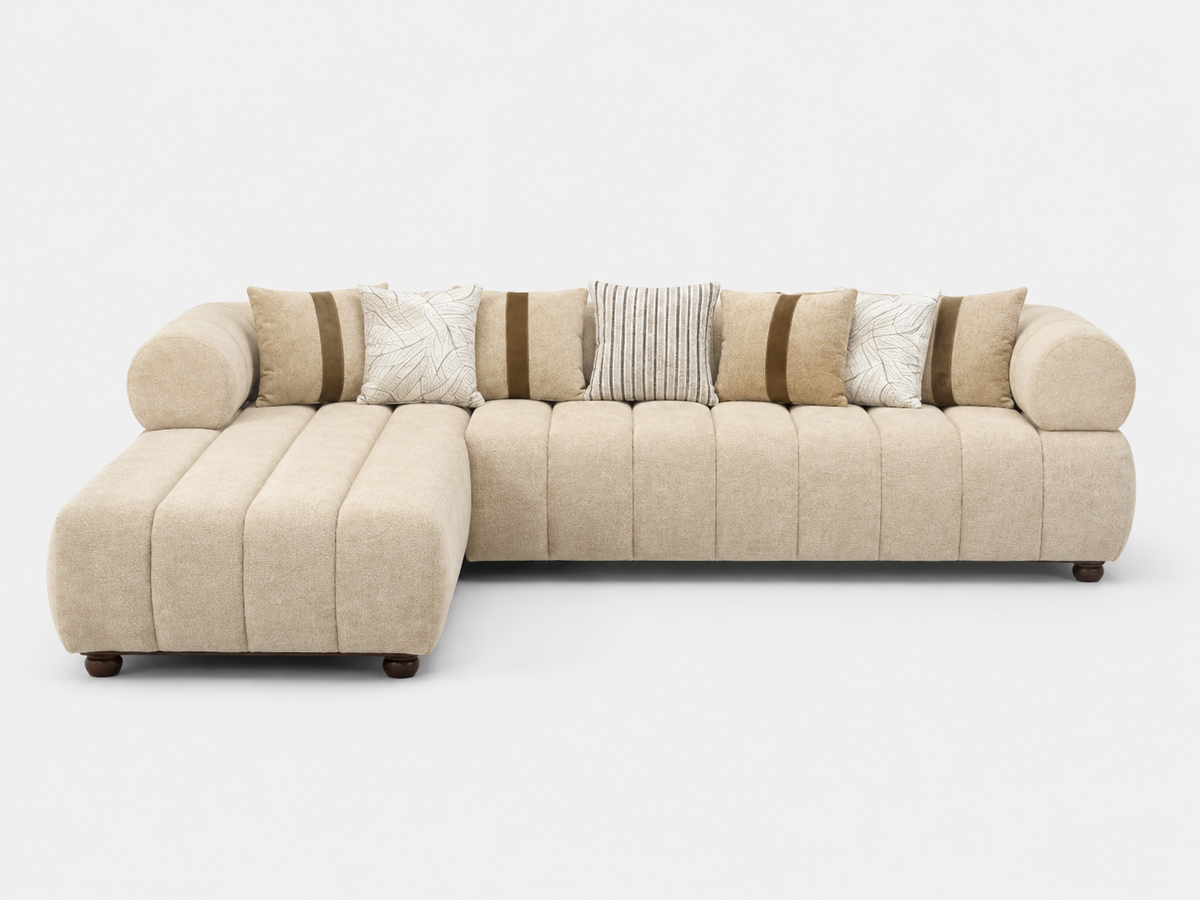 A modern L-shaped sectional sofa upholstered in a textured beige fabric, featuring a chaise lounge on the left, vertical channel tufting on the backrest, and multiple decorative throw pillows in beige, white, and striped patterns, set against a plain light gray background.